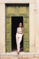 Combelle Alix Top lifestyle shot, model in fawn colourway casually propped against green doorway, front three-quarter full-length view